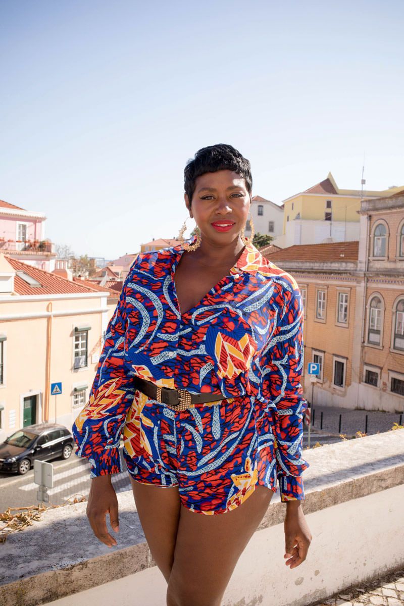 Woman in vibrant blue African print romper with gold belt poses on a sunny balcony overlooking colorful European buildings.