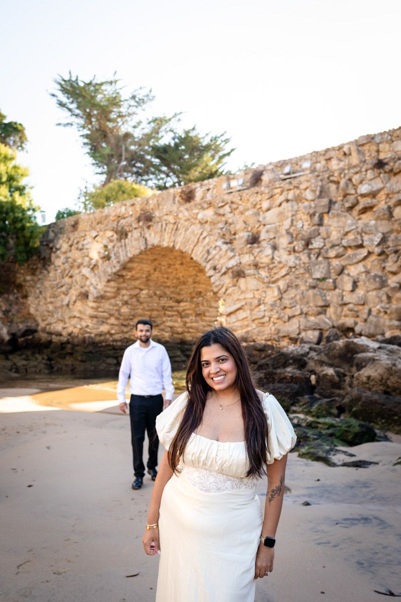 Smiling woman in white dress poses on sandy beach with stone arch and man behind her.
