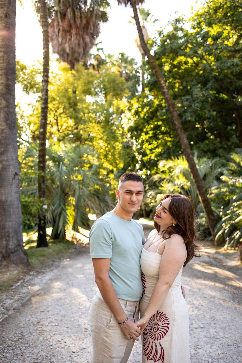 Couple holding hands on sunlit palm tree-lined path in lush tropical garden.