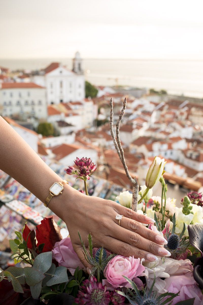 Bride's hand with engagement ring and gold watch resting on colorful bouquet with city view.