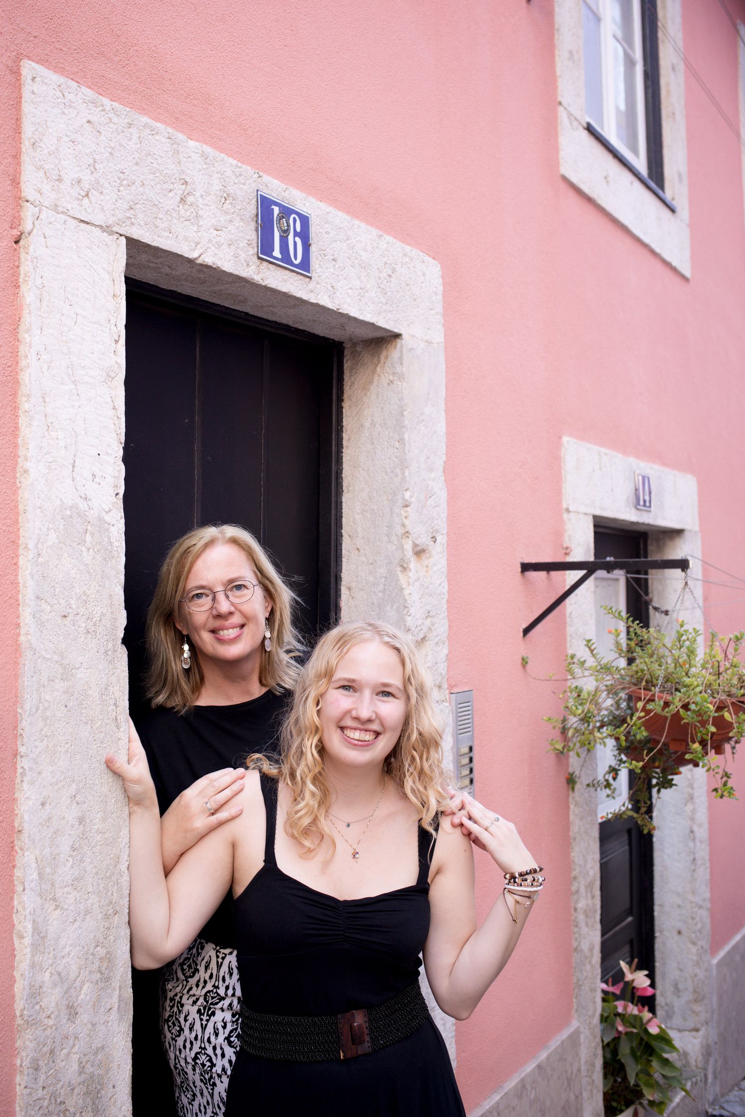 Two blonde women smiling at a pink building entrance numbered 16 in Lisbon, Portugal.