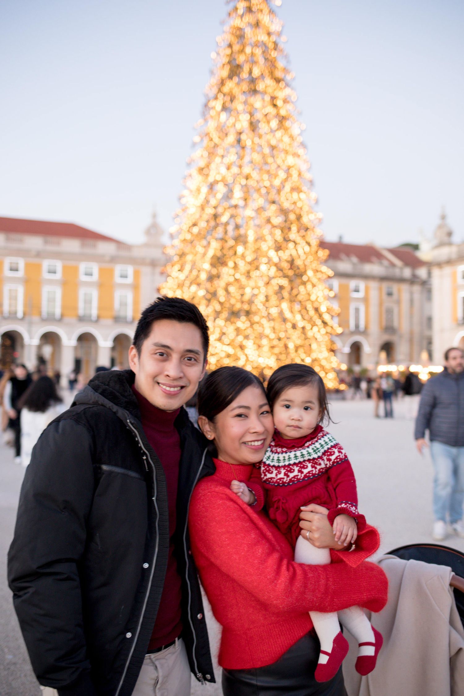 Young Asian family with toddler posing before a glowing Christmas tree at Lisbon city square.