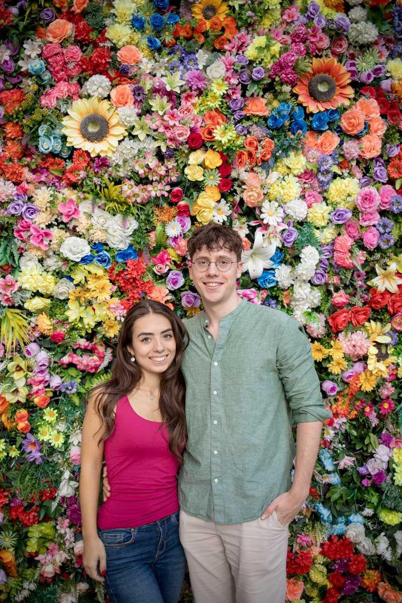 Smiling couple posing together in front of a vibrant colorful flower wall backdrop.