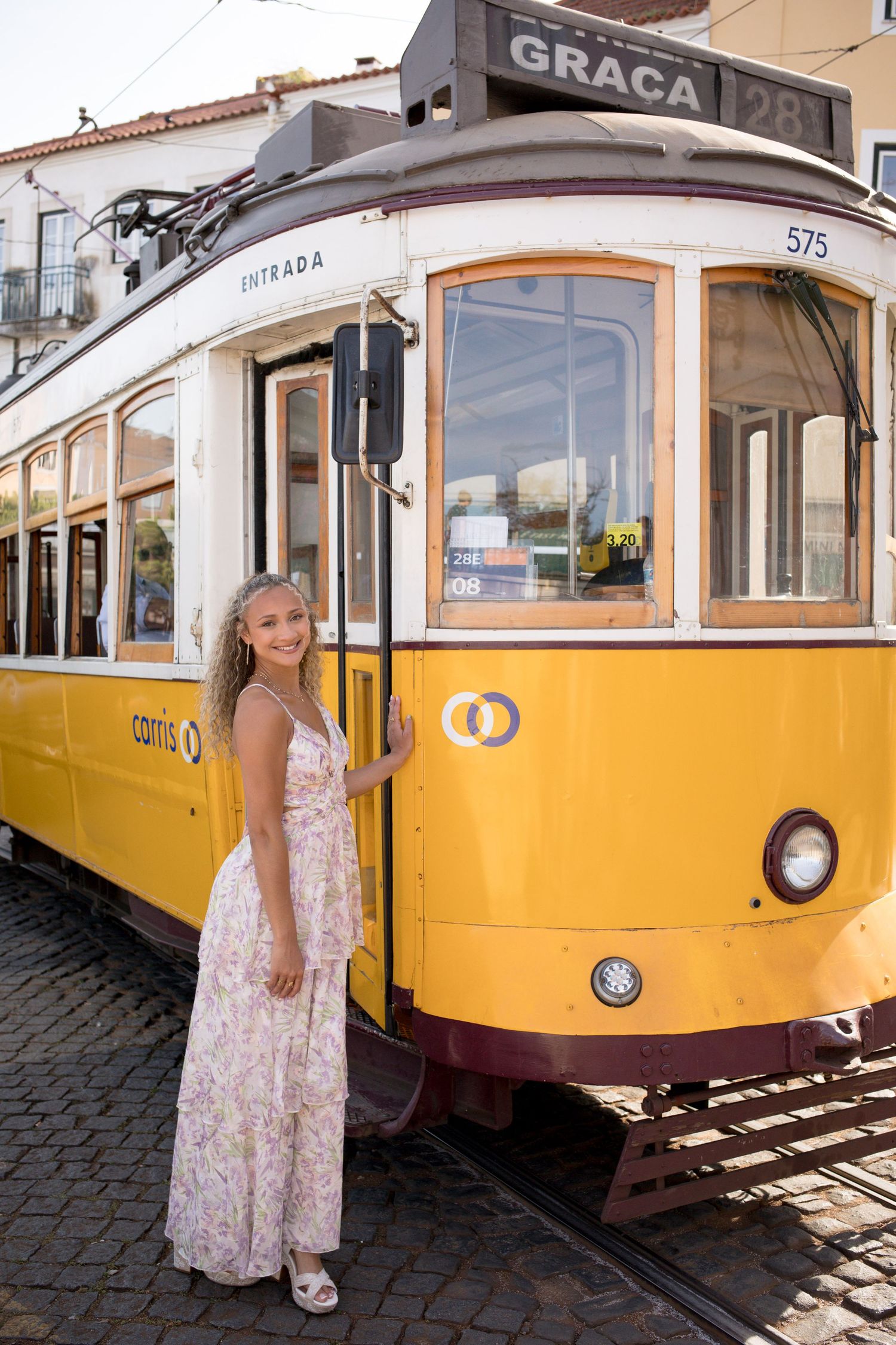 Young woman in floral dress boarding iconic yellow tram 28 on cobblestone street in Lisbon.