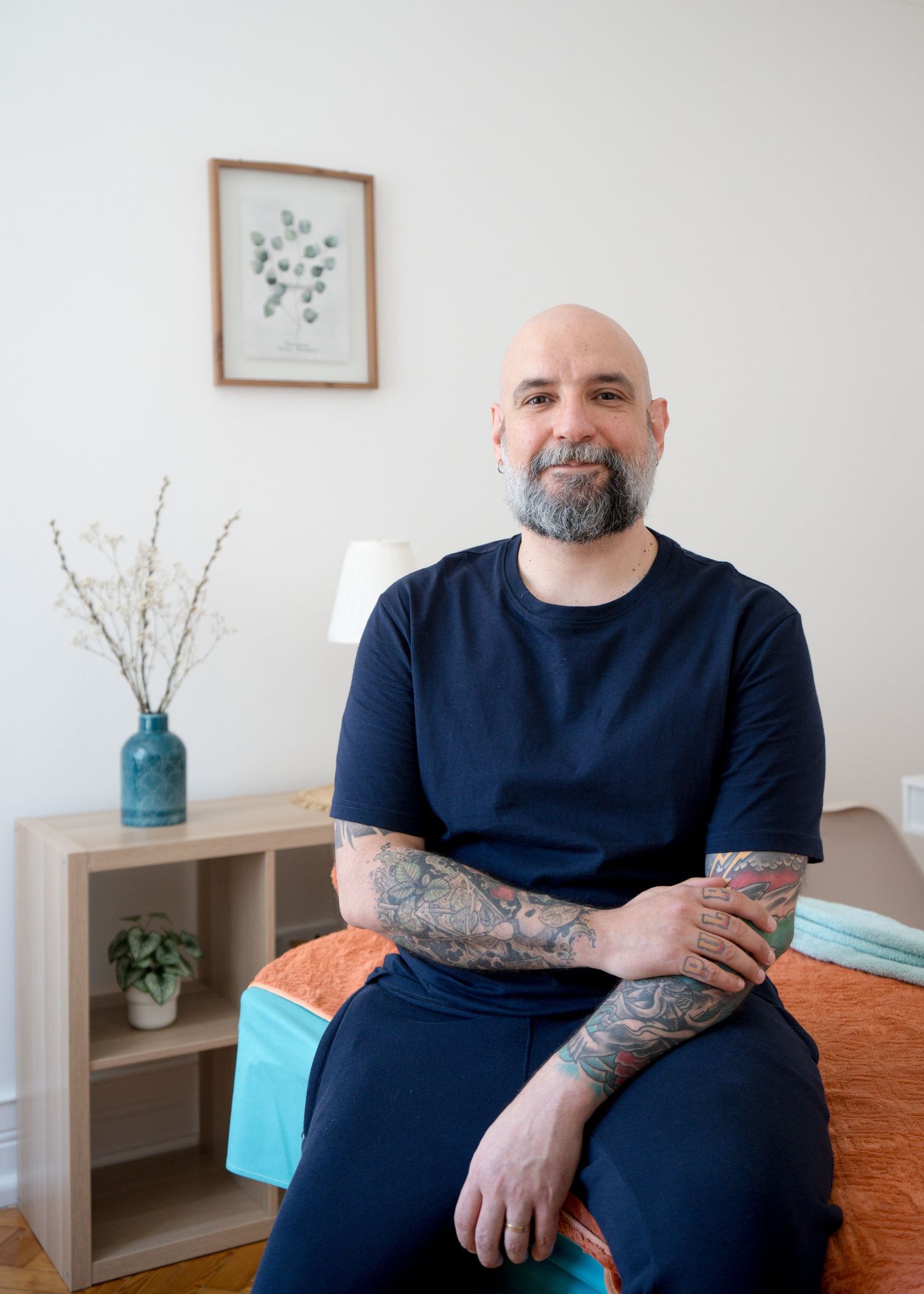 Bald man with a gray beard and tattooed forearms sits smiling on a treatment table in a bright room with a plant and framed art.
