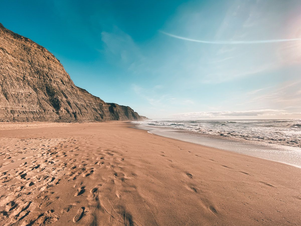 Sandy beach with footprints, dramatic cliffs, and gentle ocean waves under a bright blue sky with wispy clouds.