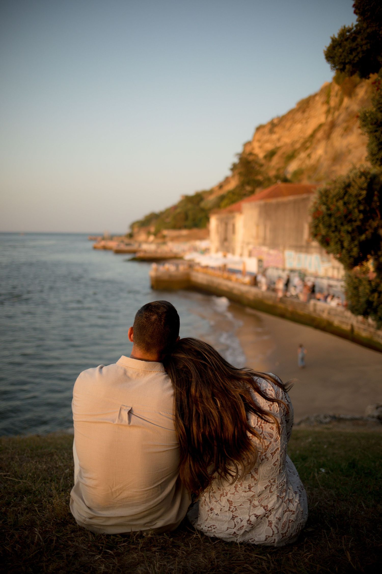 Romantic couple sitting together on a grassy cliff overlooking a serene coastal beach at golden hour sunset.
