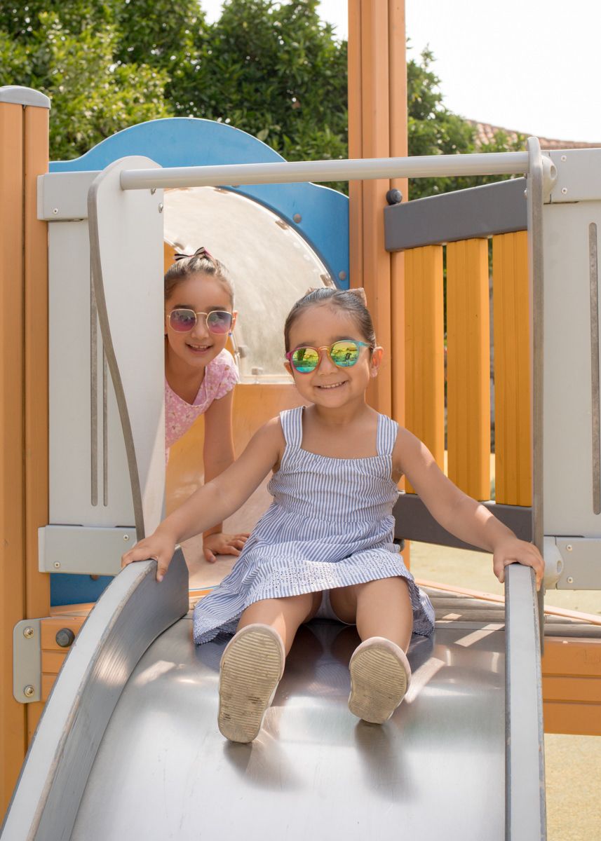 Two young girls wearing sunglasses playing on a playground slide on a sunny day.