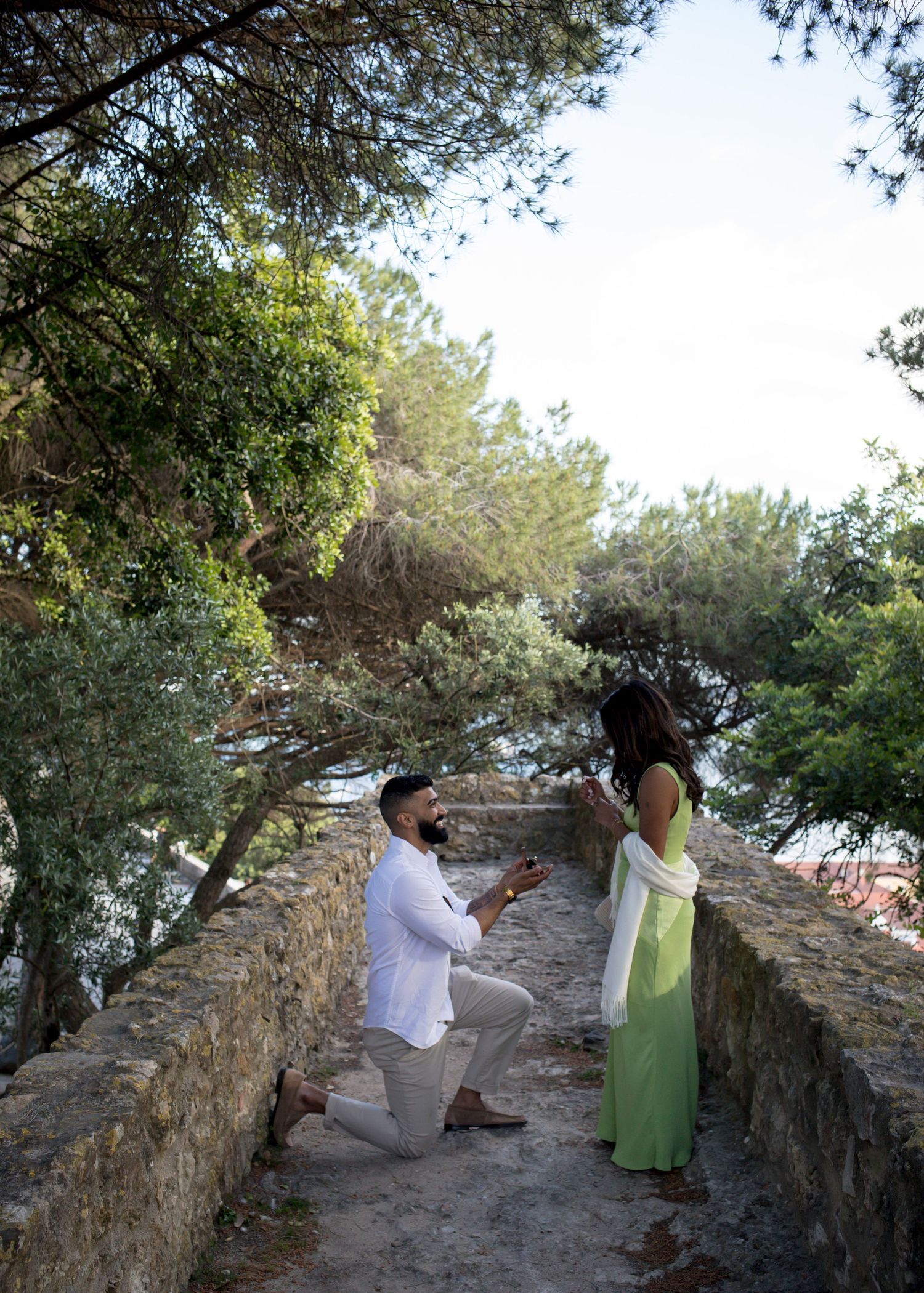 Man kneeling on stone wall proposing to woman in green dress surrounded by lush trees at scenic outdoor location.