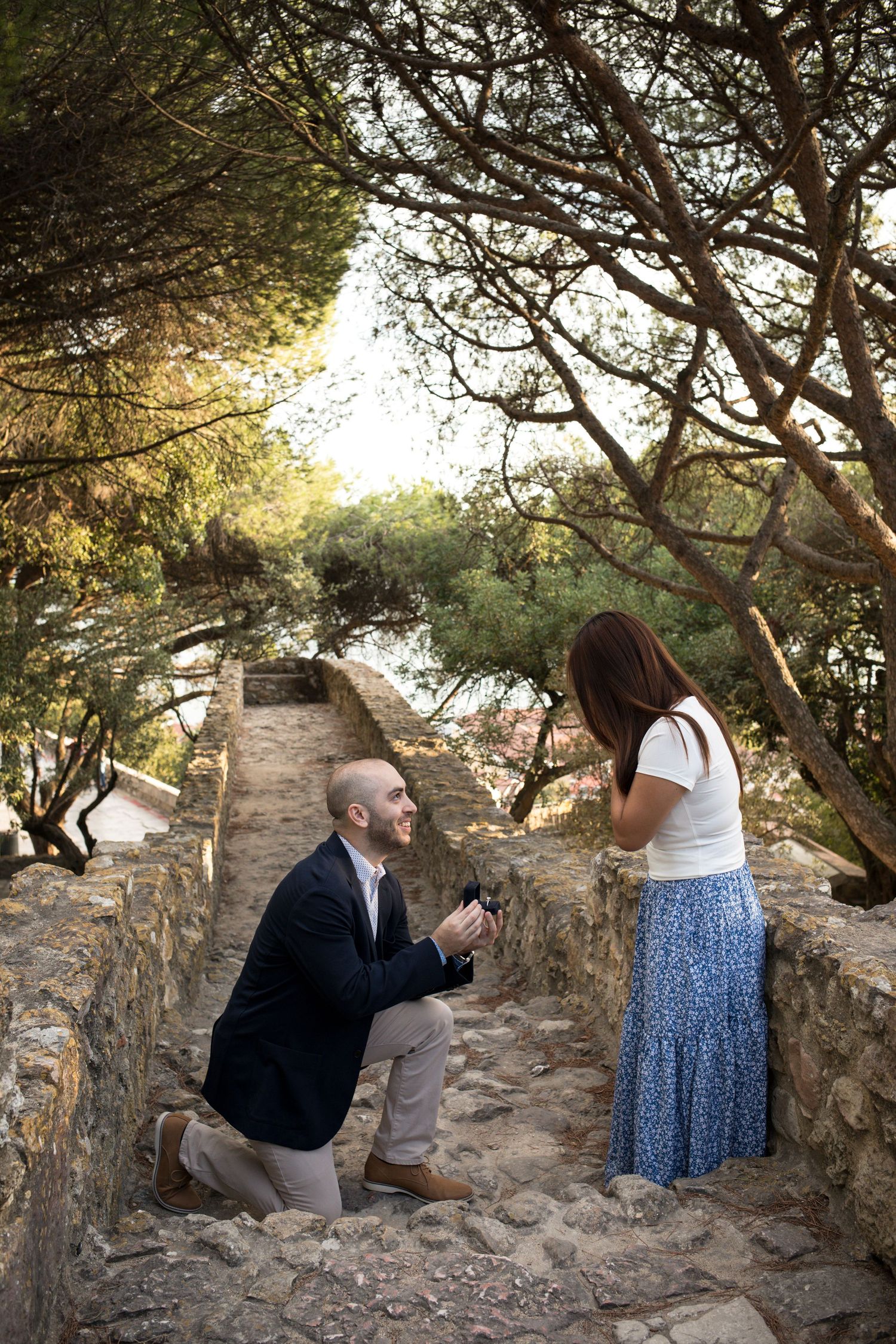 Man kneeling on stone path proposing to woman in blue skirt surrounded by lush trees at golden hour.