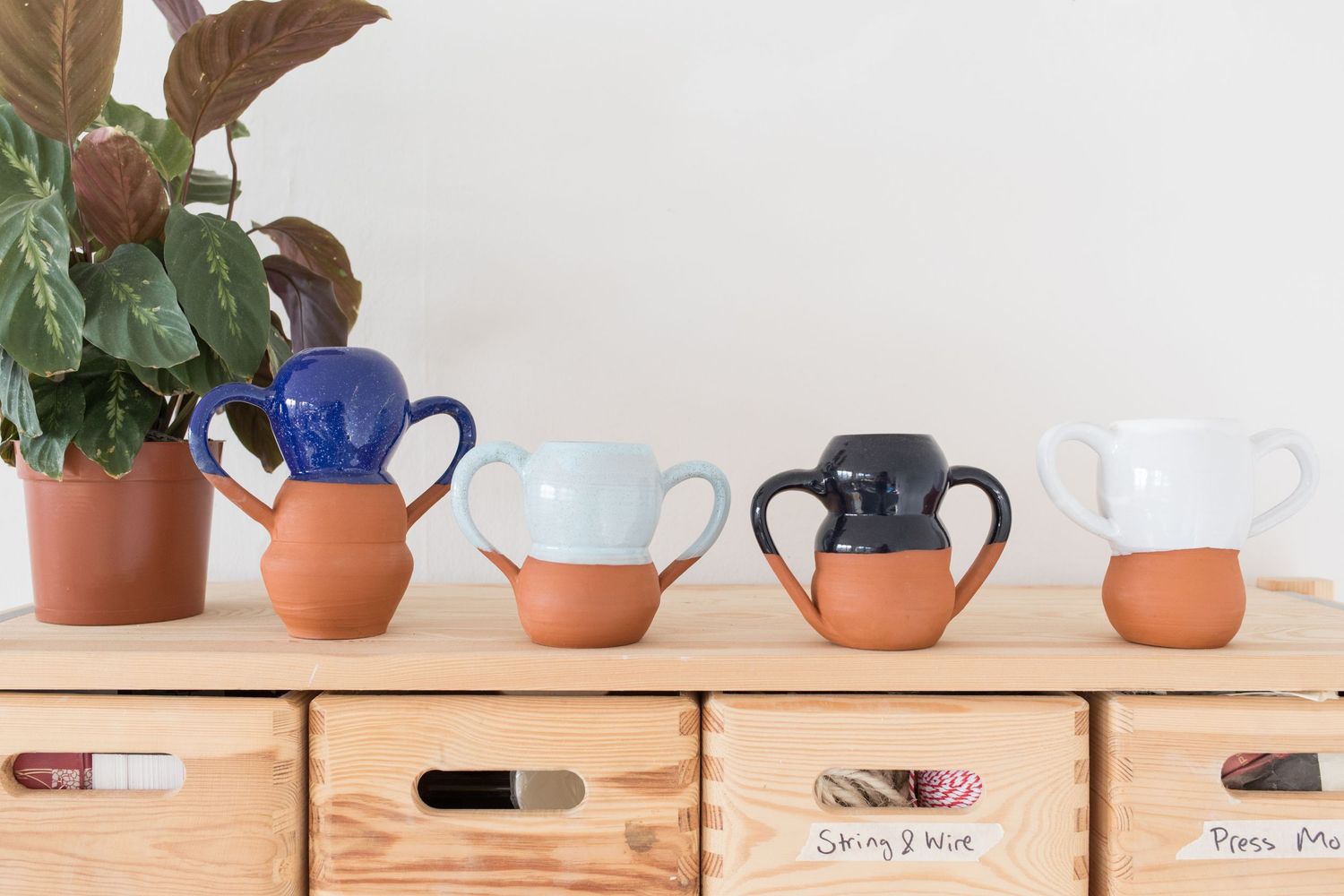 Four handmade two-handled ceramic mugs with terracotta bases and blue, white, and black glazes on a wooden shelf beside a leafy plant.