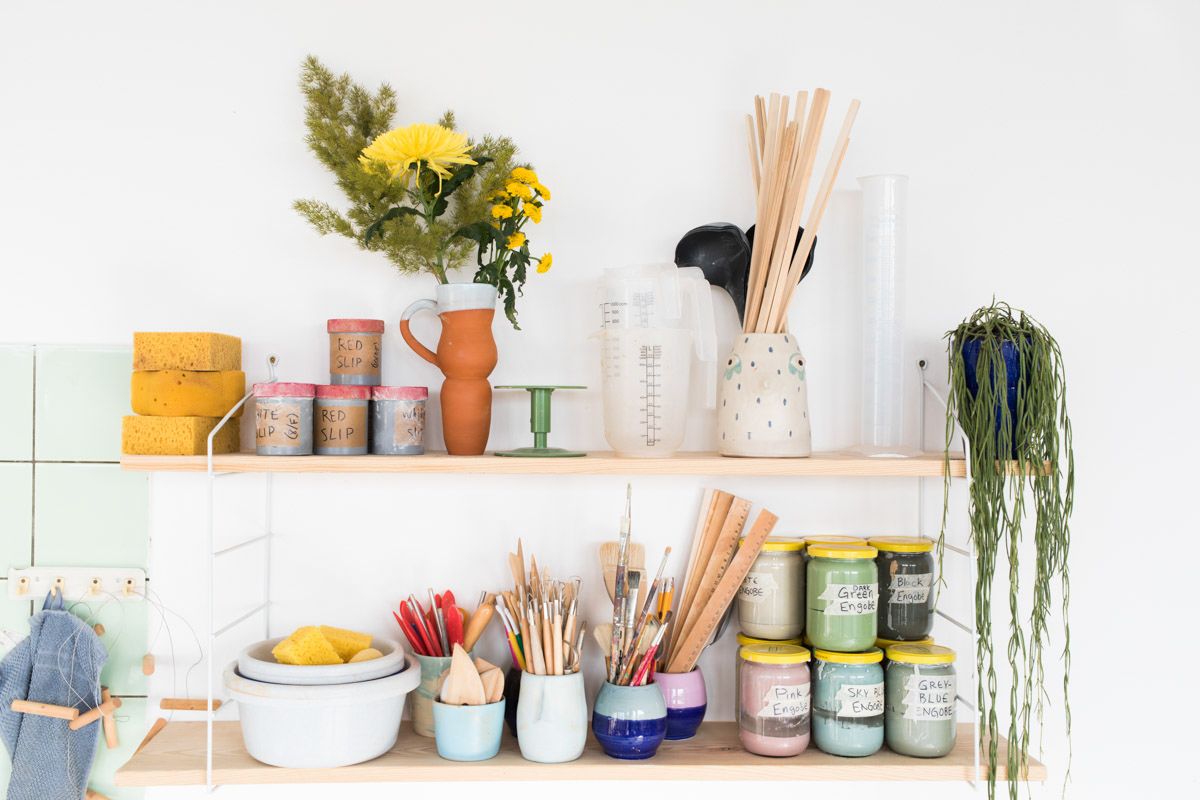 Colorful craft supplies and paint jars displayed on wooden shelves with flowers and brushes in a bright studio.