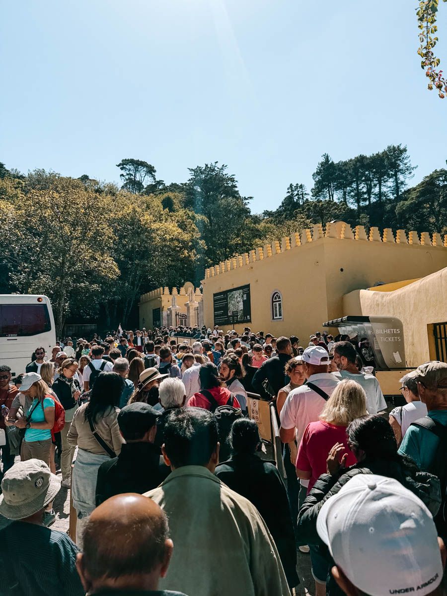 Large crowd of tourists queuing outside a yellow historic palace entrance surrounded by lush trees on a sunny day.