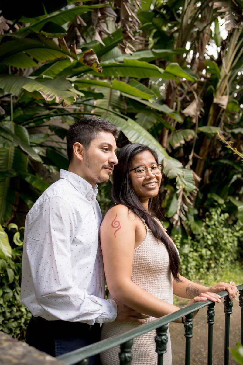 Couple smiling together on a balcony surrounded by lush tropical greenery, woman with a red tattoo wearing a beige dress.