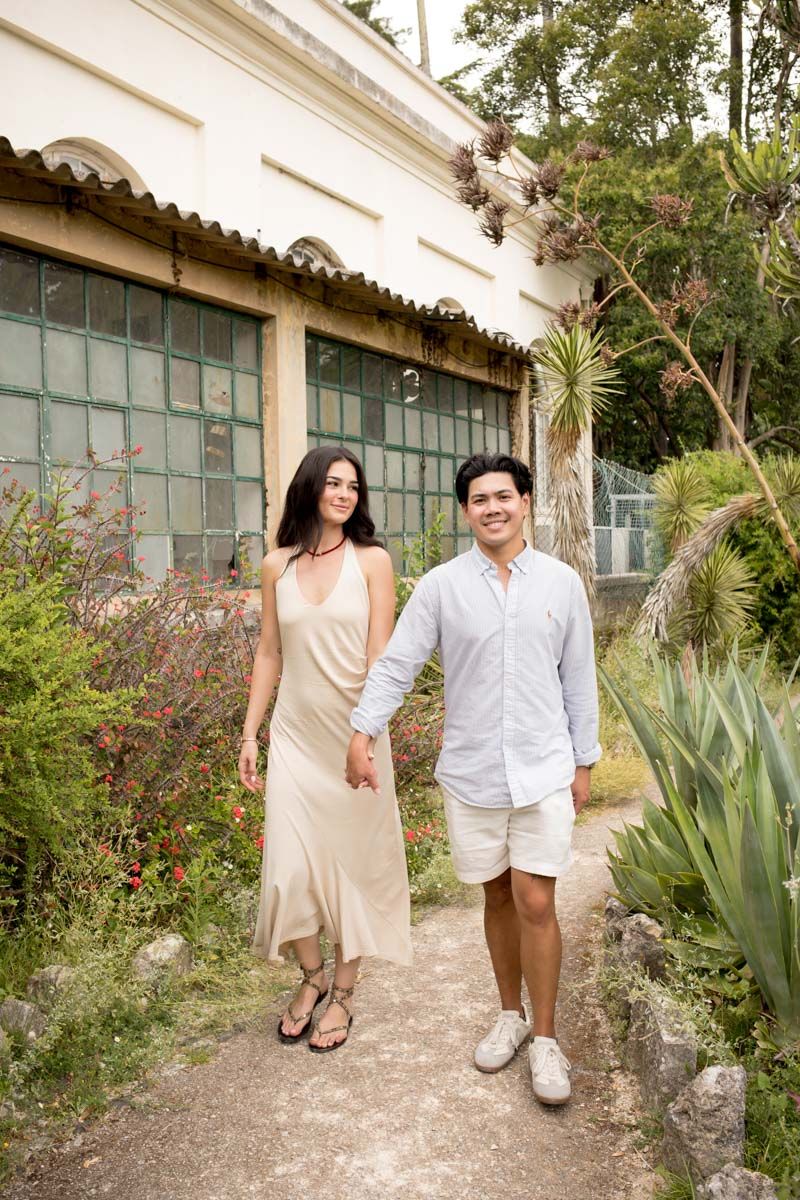 A couple holds hands walking through a lush garden path beside a white building with green windows.