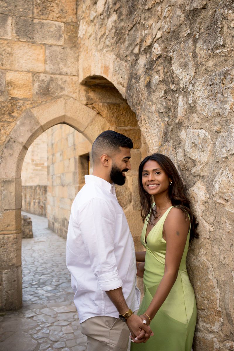 Couple holding hands near a stone archway, woman in green dress smiling at camera.