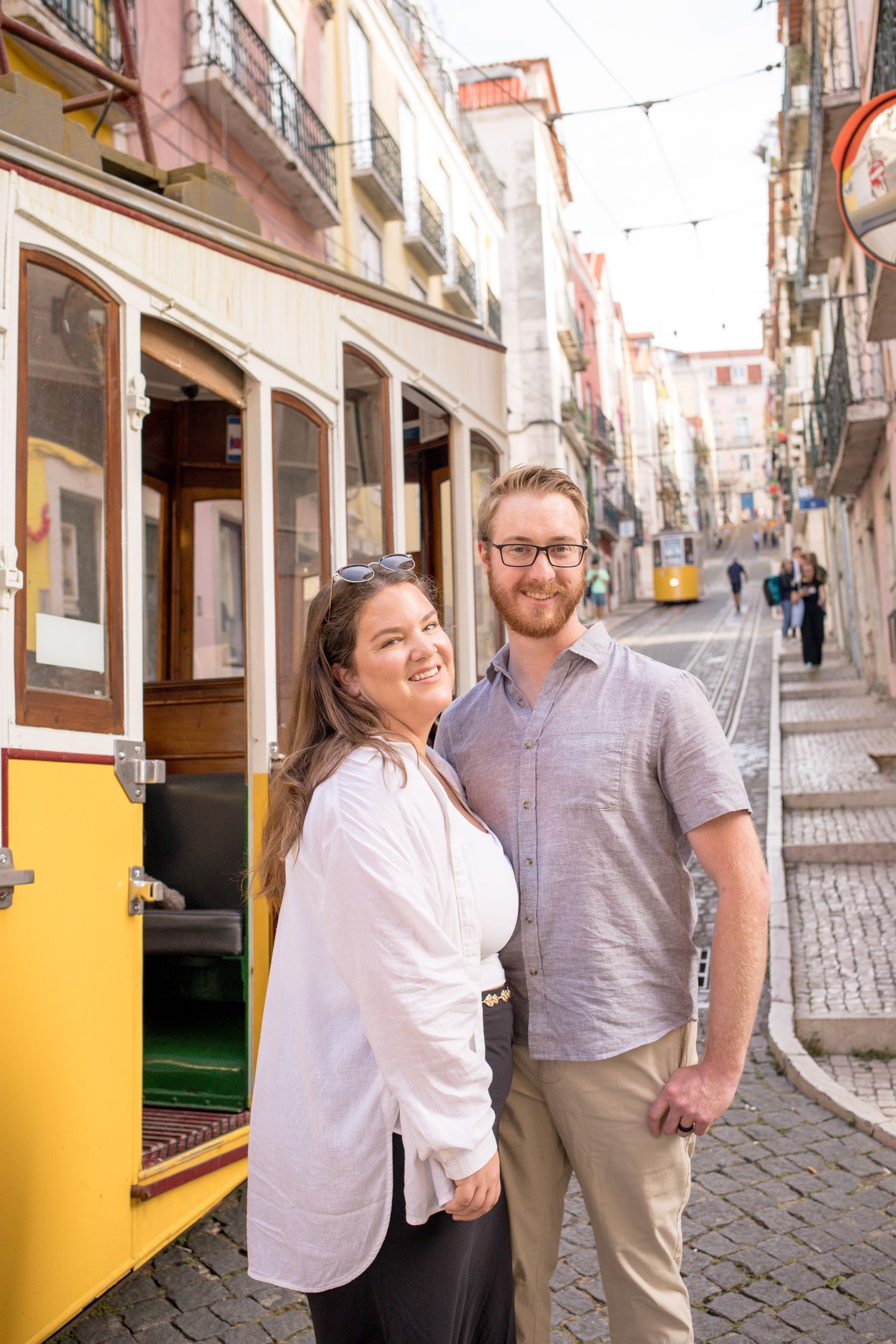 Smiling couple posing beside a classic yellow Lisbon tram on a cobblestone street in Portugal.