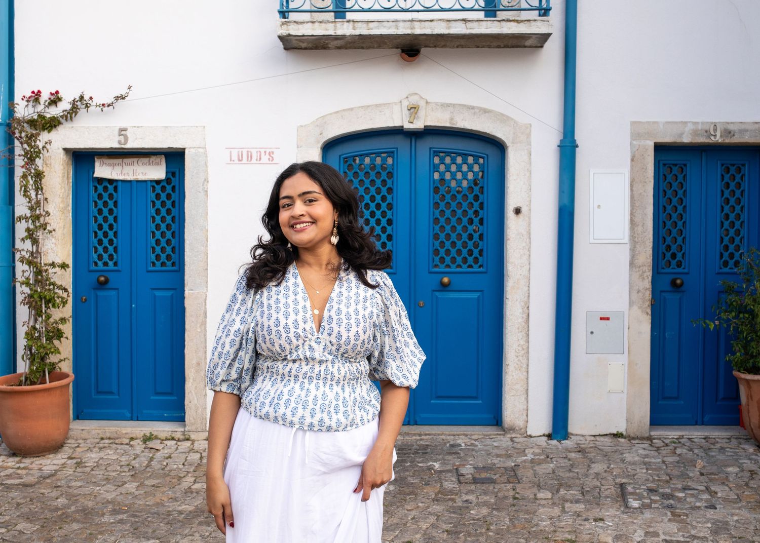 Smiling woman in a printed blouse standing before charming blue doors in a Portuguese town.