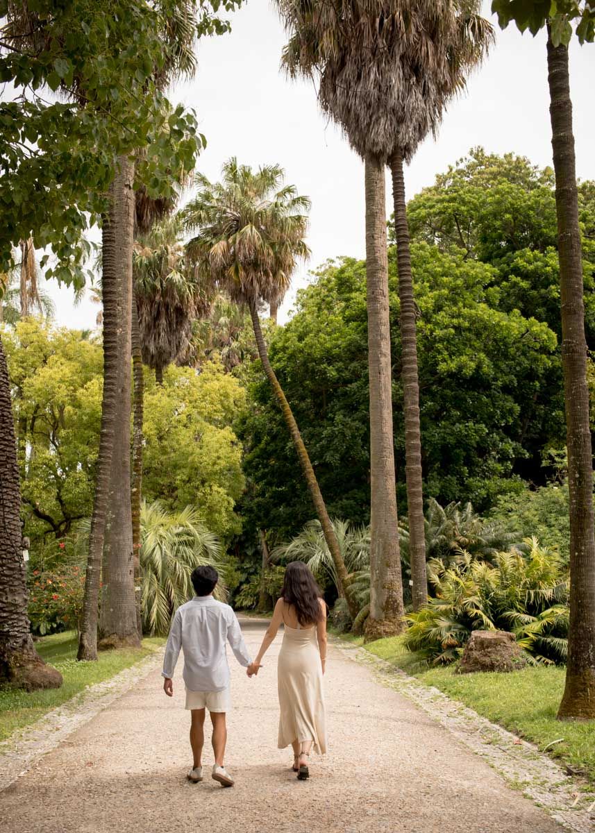 Couple holding hands walking down a palm tree-lined garden path surrounded by lush tropical greenery.