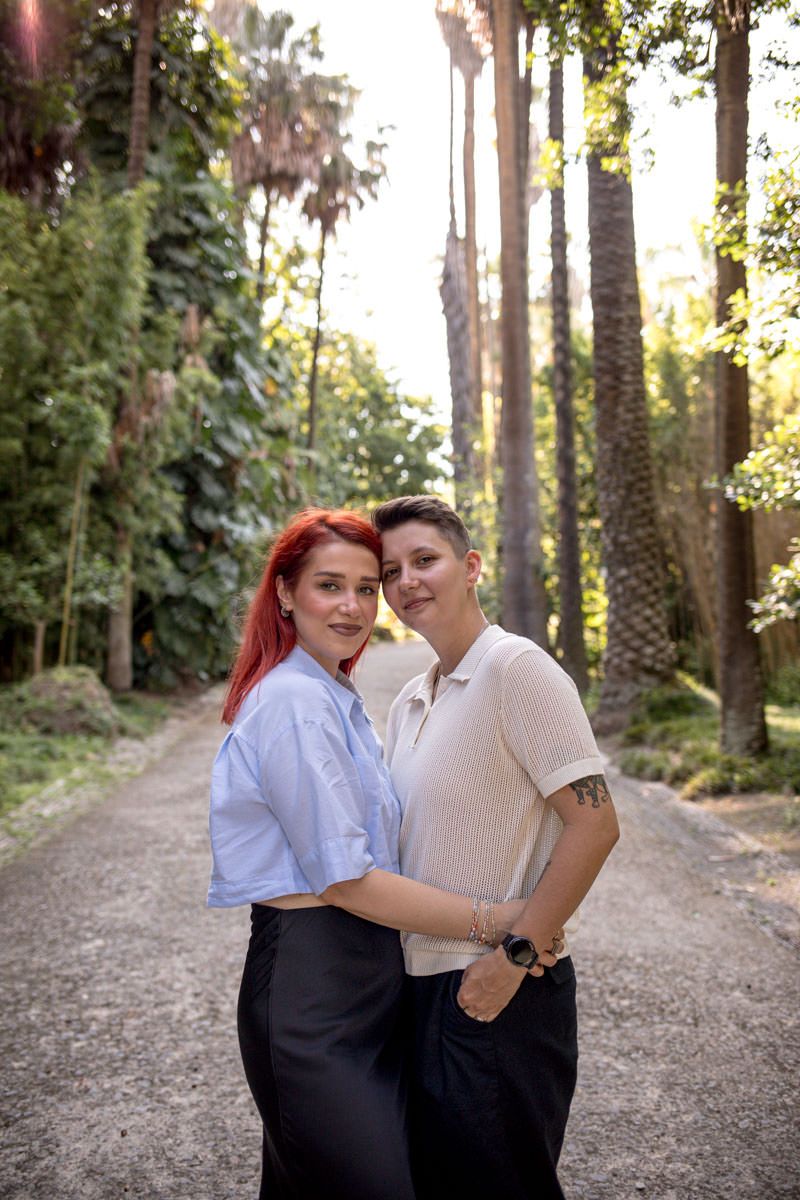 Two women embracing on a sunlit forest path surrounded by tall palm trees, smiling warmly at the camera.