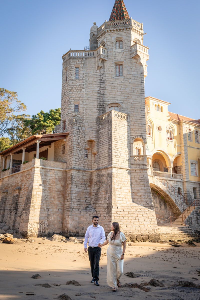 Couple holding hands on sandy beach in front of historic stone castle tower in Cascais, Portugal.