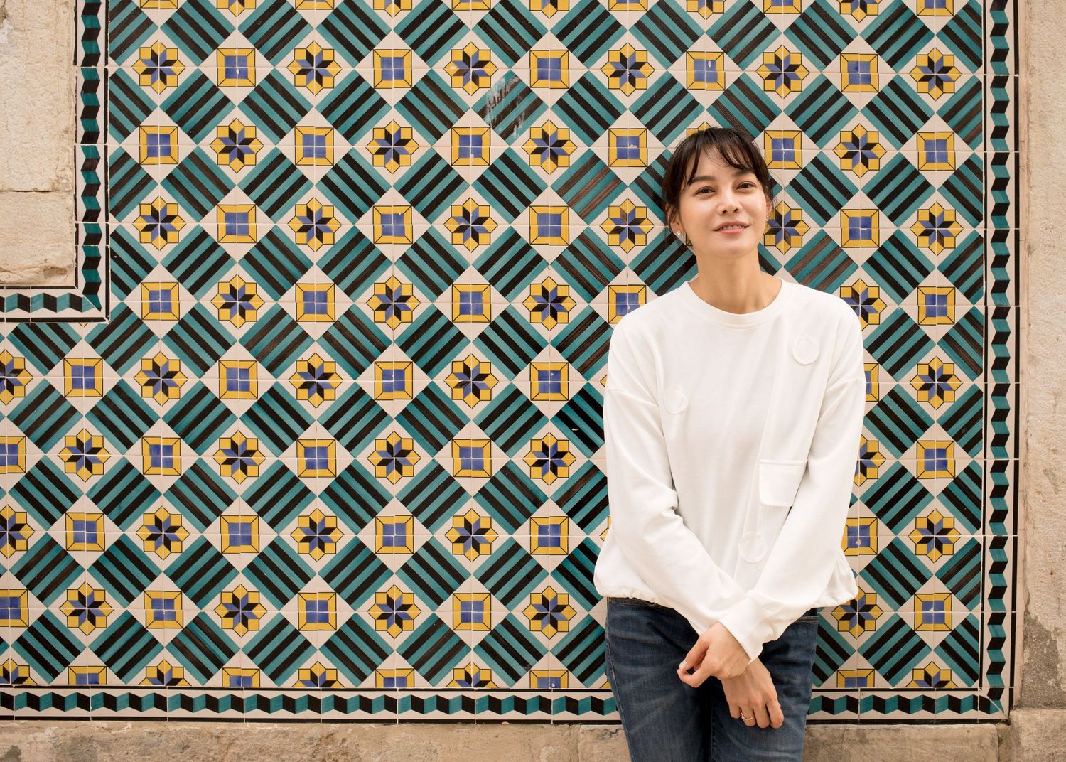 Asian woman in white sweater sitting against colorful geometric tiled wall smiling.