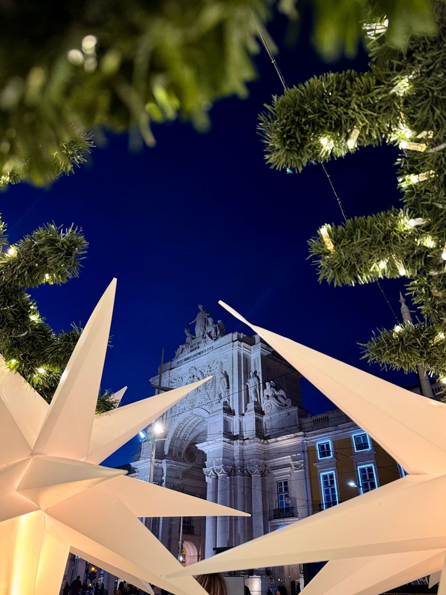 Christmas decorations with star ornaments and lit garlands frame a historic triumphal arch at night in Lisbon, Portugal.