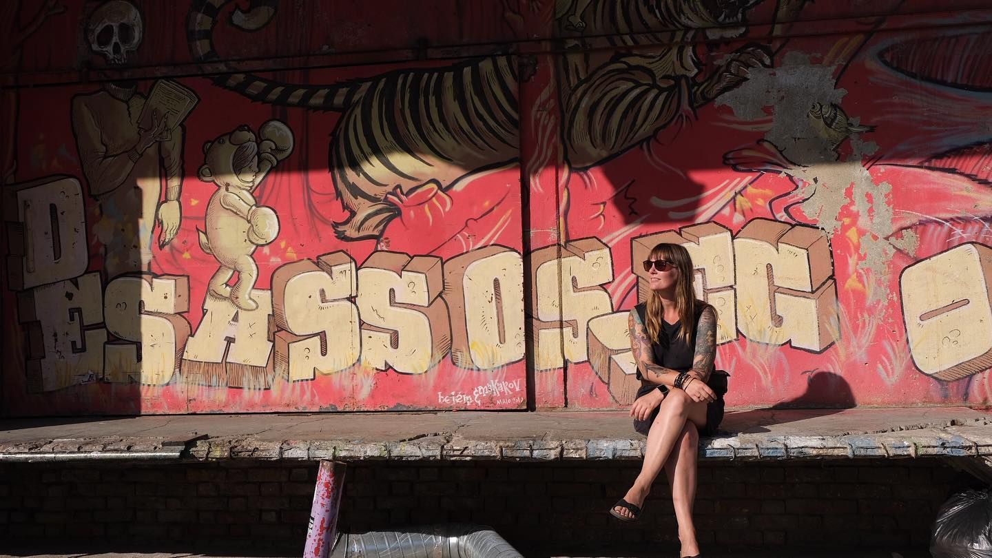 Woman in black outfit sitting before vibrant graffiti mural with tiger artwork on urban wall.