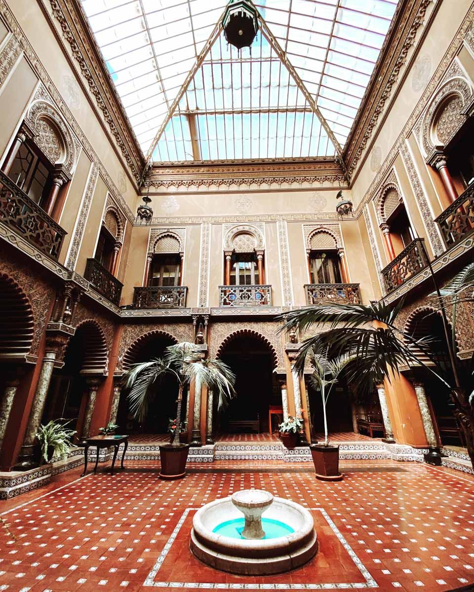Ornate Moroccan riad courtyard with fountain, palm trees, arched galleries, and a stunning glass skylight ceiling.