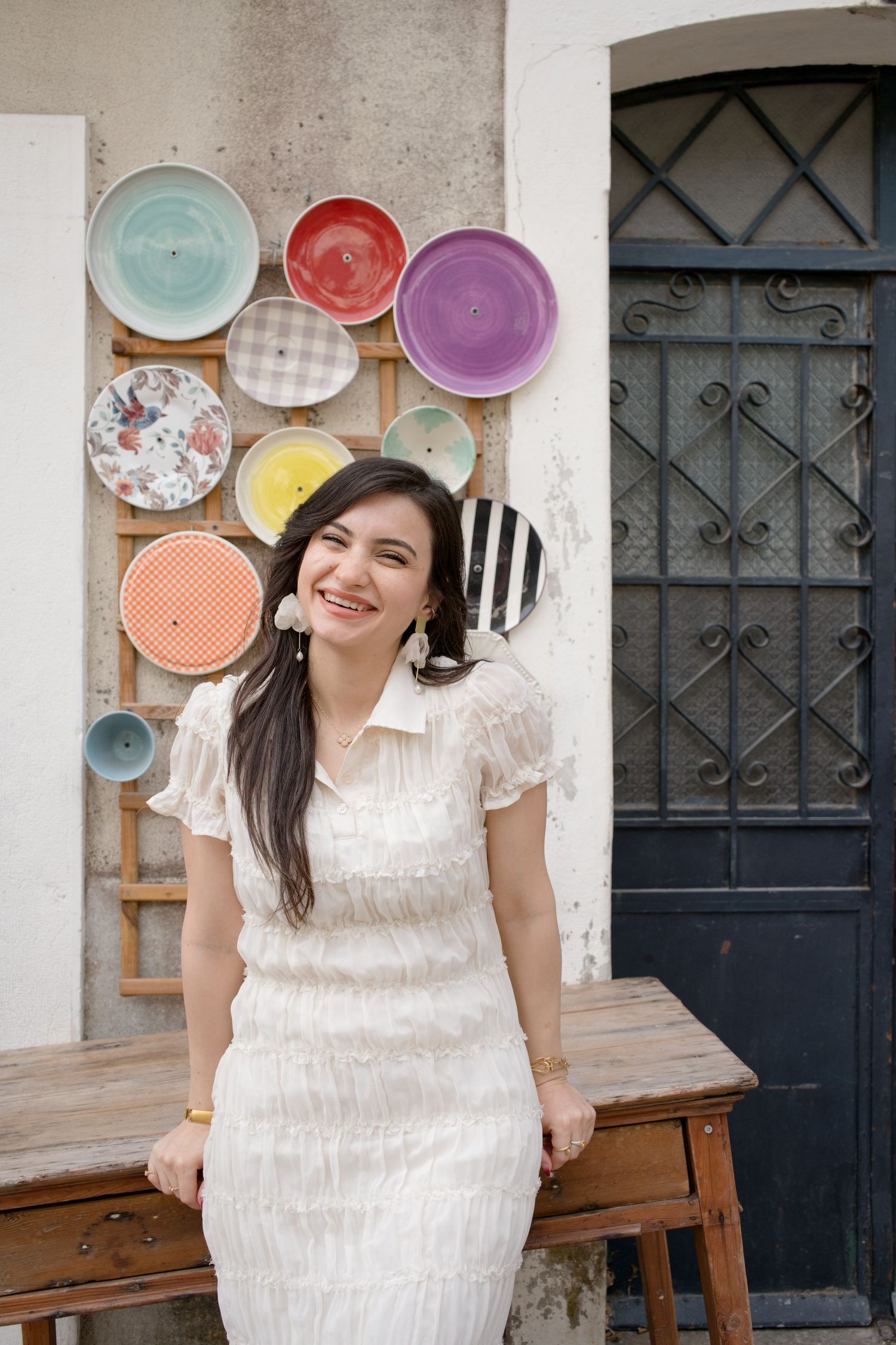Smiling woman in a white dress leans on a rustic wooden table in front of a wall of colorful decorative plates and a dark wrought-iron door.