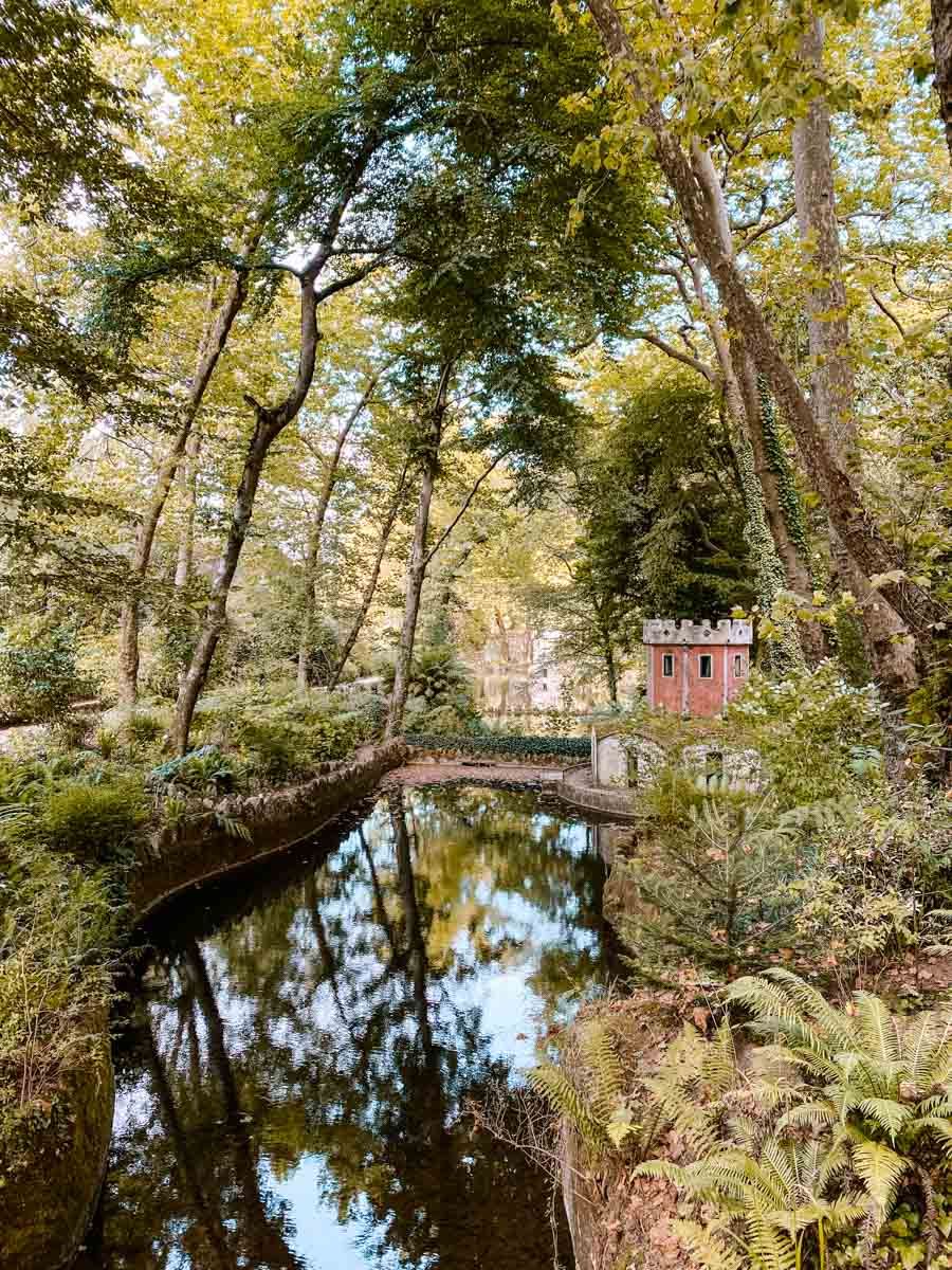 A serene forest canal with stone bridge and red brick tower reflected in still water, surrounded by lush green trees.