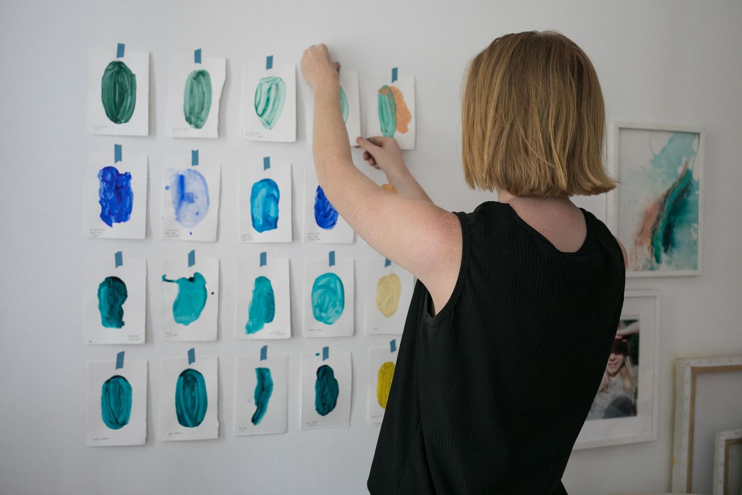 Woman in a black top pins a small watercolor swatch to a white wall covered with rows of blue and green paint samples.