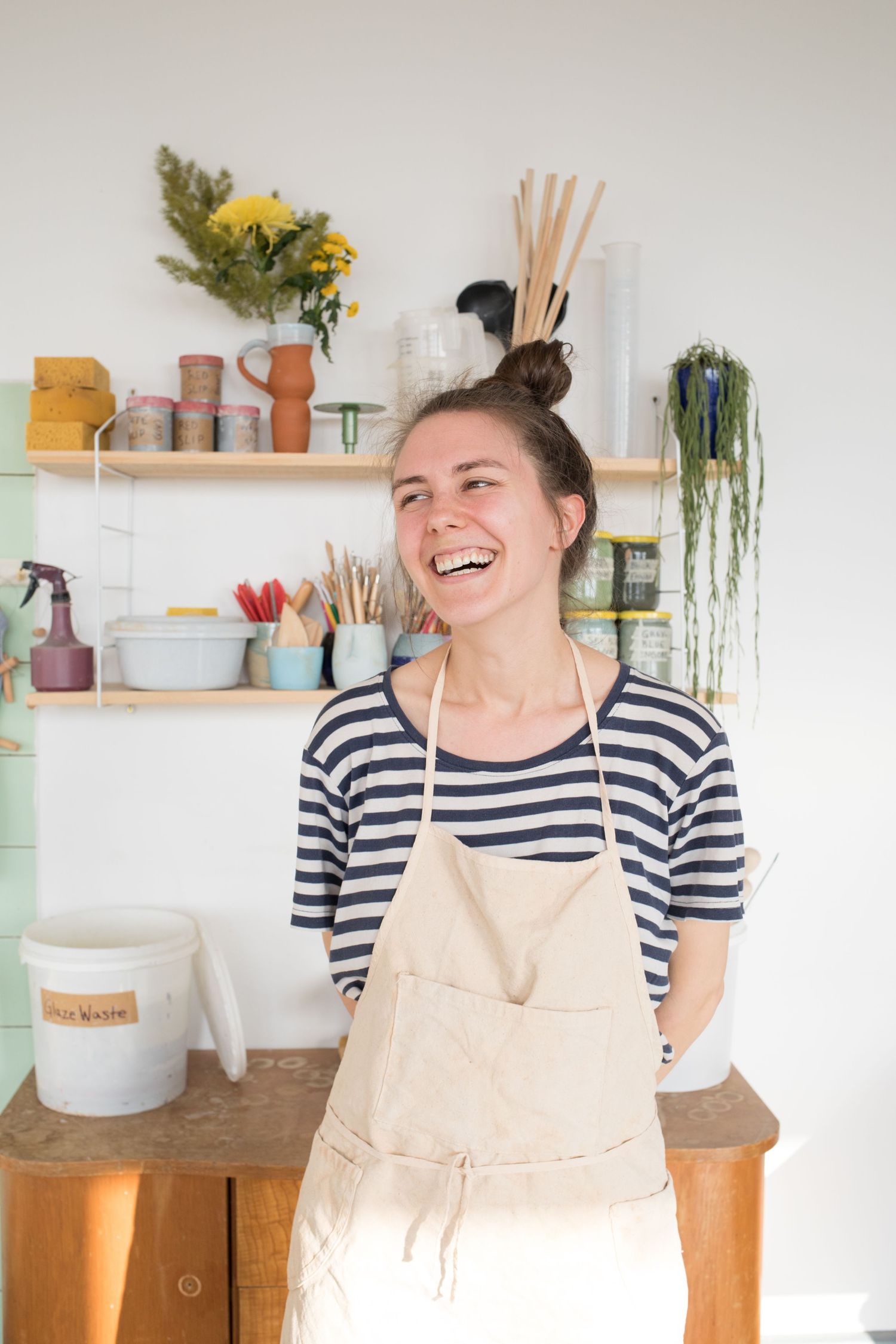 Smiling woman in a striped shirt and linen apron laughs in a bright pottery studio with shelves of jars, brushes, and plants.