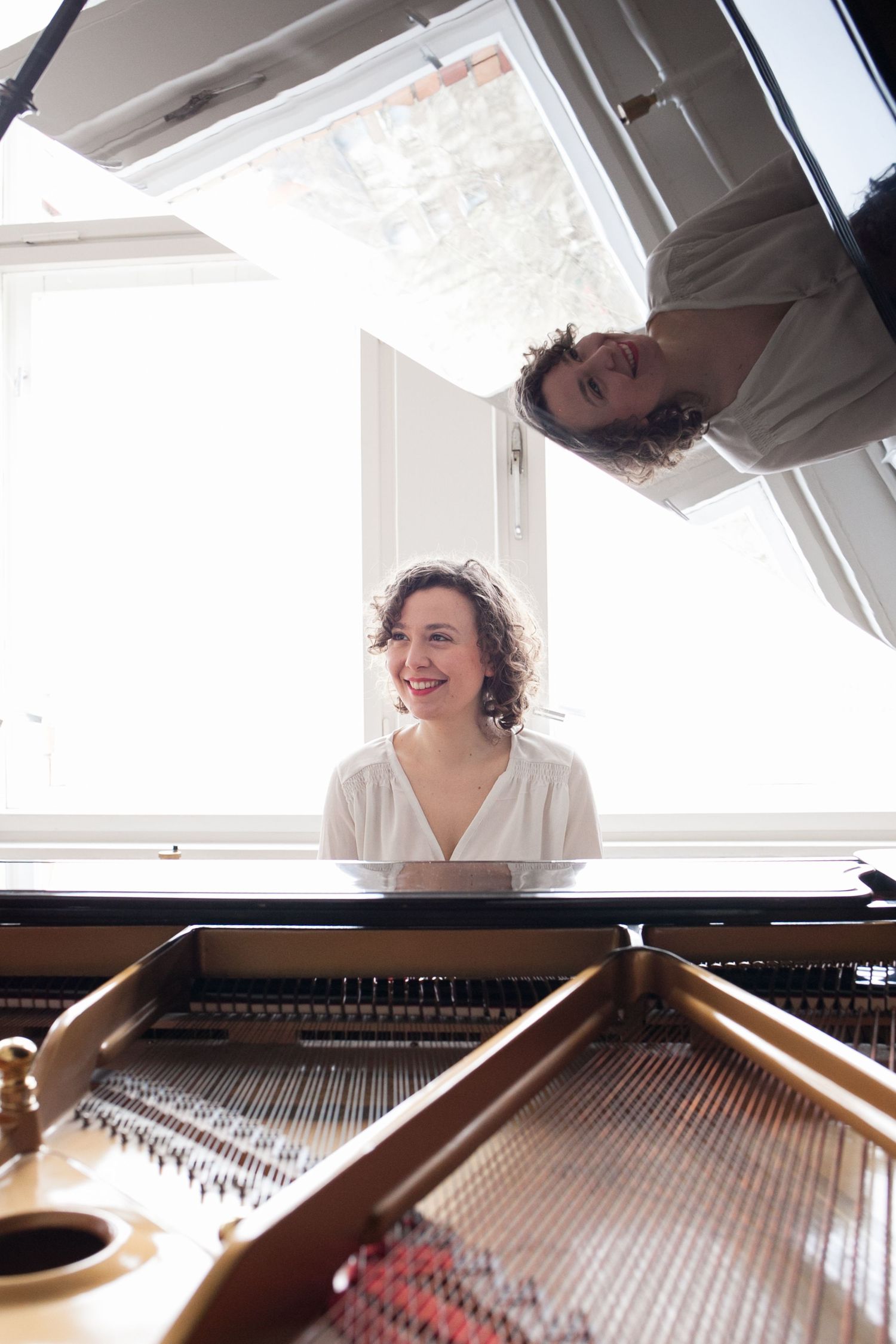 Smiling woman with curly hair sits at an open grand piano in a bright room, her reflection visible in the raised lid above.