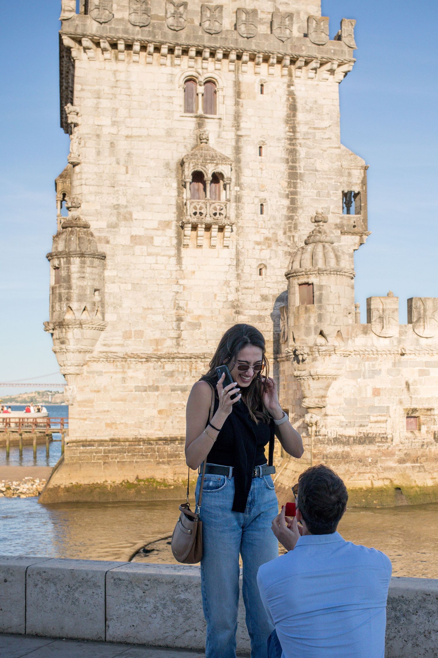 Couple near Torre de Belem in Lisbon, woman on phone, man kneeling, historic tower by the Tagus River.