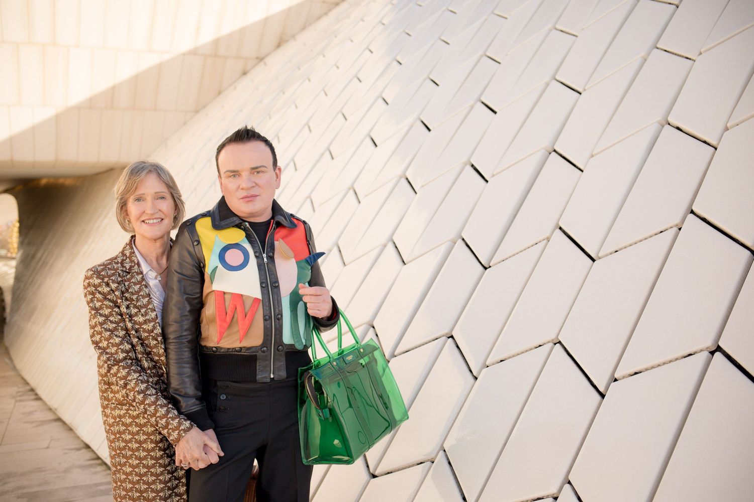 Two people smiling and posing together outdoors near a modern geometric white wall architecture.