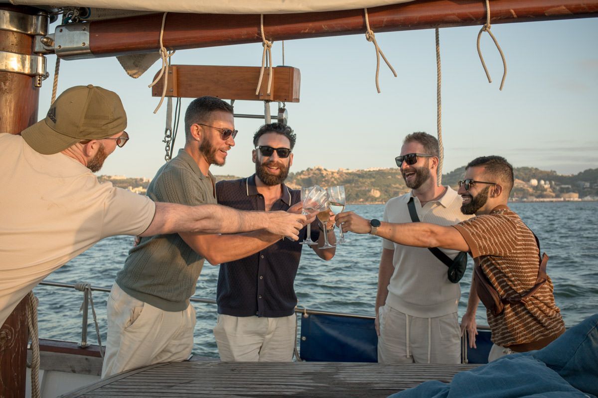 Group of five men toasting drinks on a boat deck with ocean views at sunset.