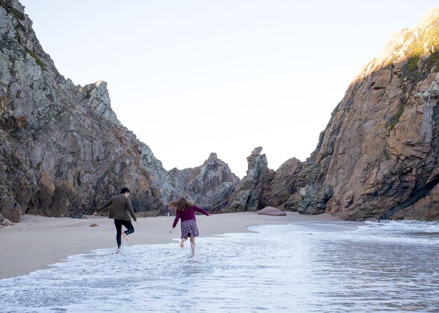 Two people running playfully along a sandy beach surrounded by dramatic rocky cliffs.