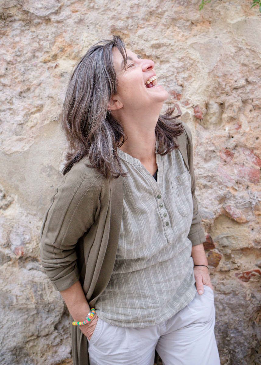 Woman with gray-streaked hair laughs joyfully while leaning against rustic stone wall outdoors.