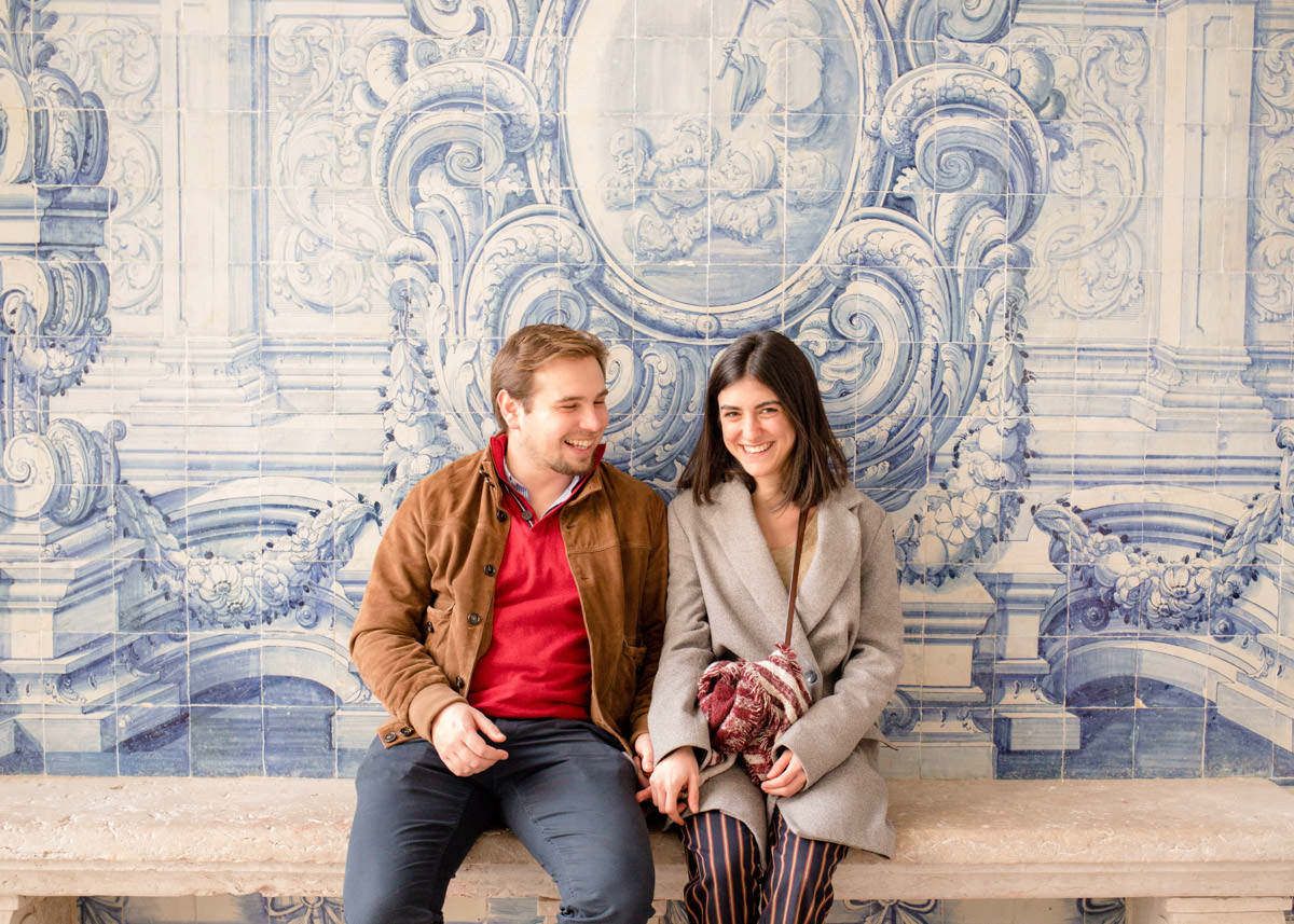 Couple sitting together on a bench in front of ornate blue and white Portuguese azulejo tile wall.