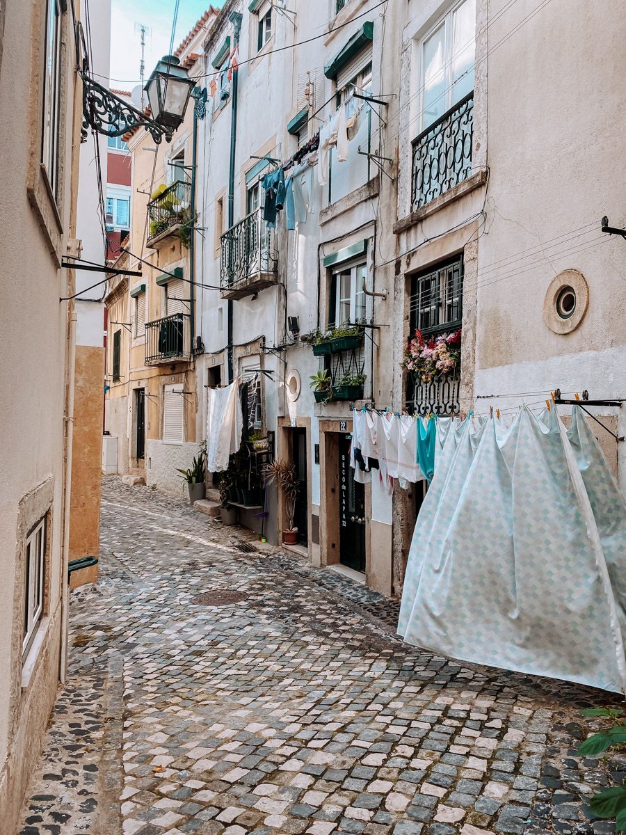 Narrow cobblestone alley in Lisbon with laundry hanging between old pastel buildings on a sunny day.