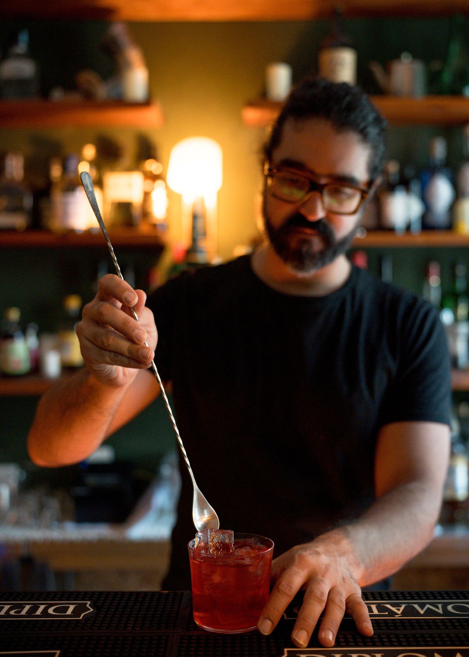 Bartender with glasses and a beard stirs a red cocktail with a long bar spoon at a bar with shelves of bottles behind him.