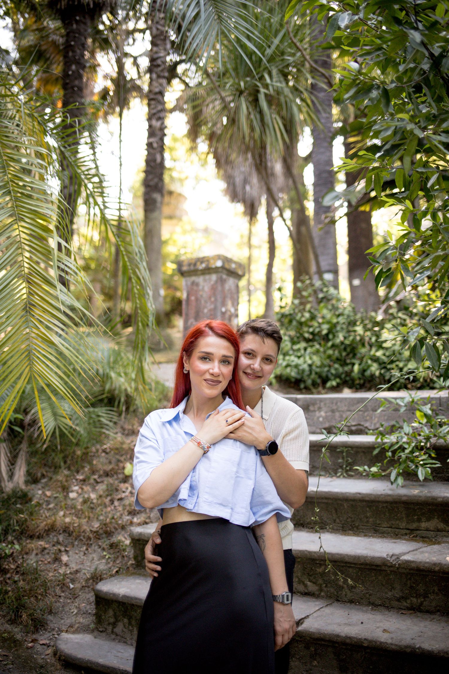 Couple embraces on stone steps surrounded by palm trees, woman with vibrant red hair in a sunlit garden.