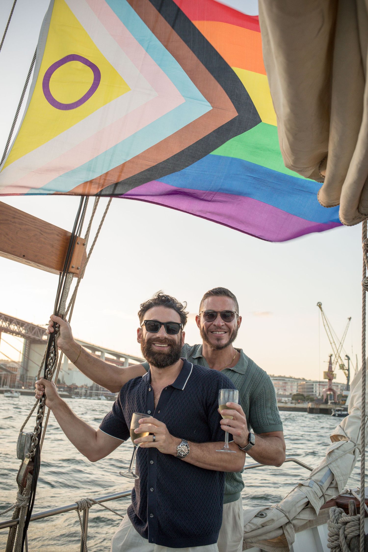 Two men holding champagne glasses on a sailboat with a Progress Pride flag, celebrating on the water at sunset.