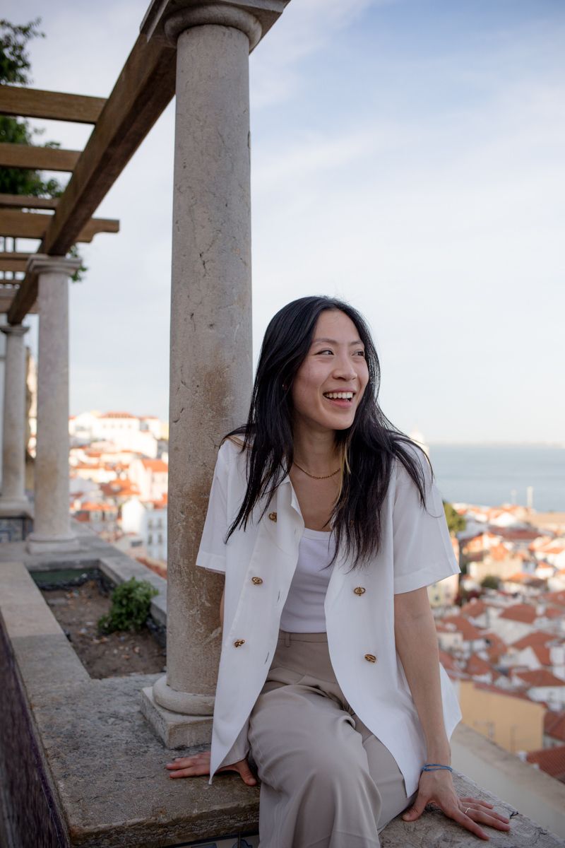 Smiling Asian woman in white blazer sits on stone ledge with Lisbon cityscape and sea behind her.