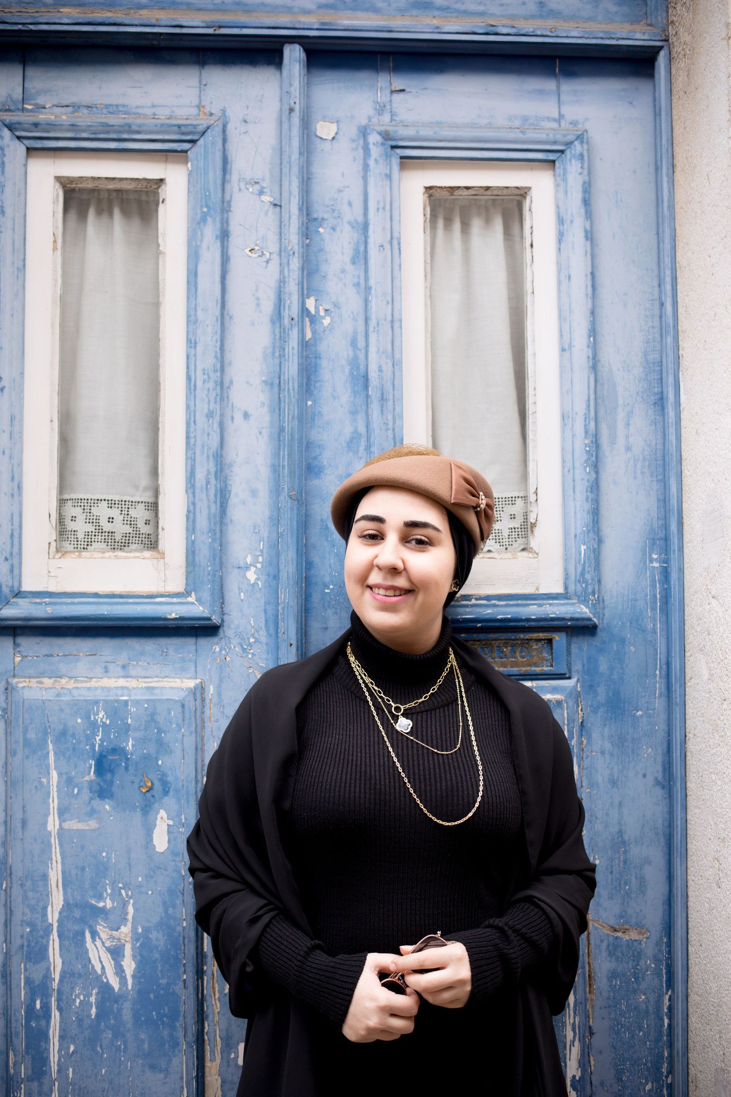 Smiling woman in black outfit and brown beret standing before rustic blue door.