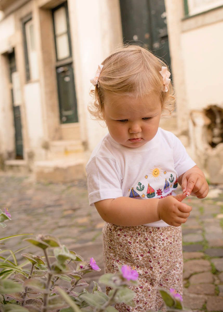 Toddler girl with blonde hair and bow clips examines flowers on the streets of Alfama in Lisbon in a white graphic tee and floral skirt.