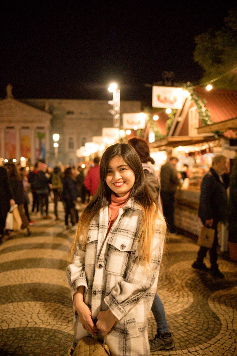 Young woman in plaid coat smiling at a festive night market with string lights and crowd.