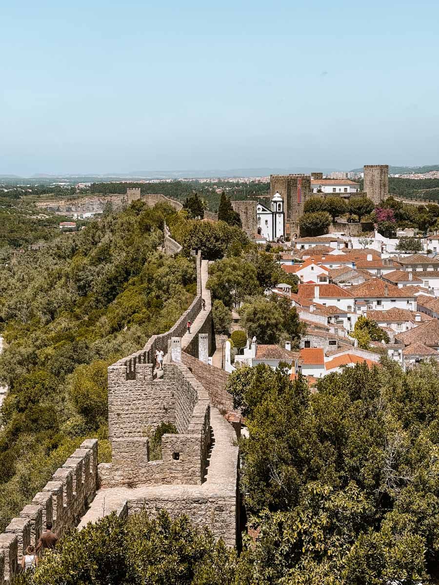 Aerial view of medieval Óbidos castle walls and whitewashed village with terracotta rooftops surrounded by lush trees, Portugal.