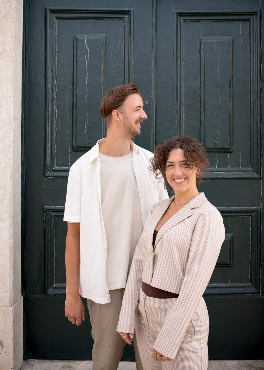 A smiling couple in neutral tones posing together in front of a dark green door.